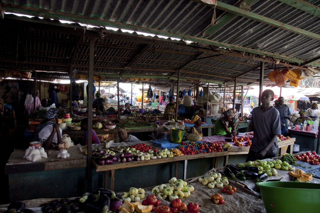 Vendors at wooden tables piled with aubergines, tomatoes and squash, under a corrugated iron roof.