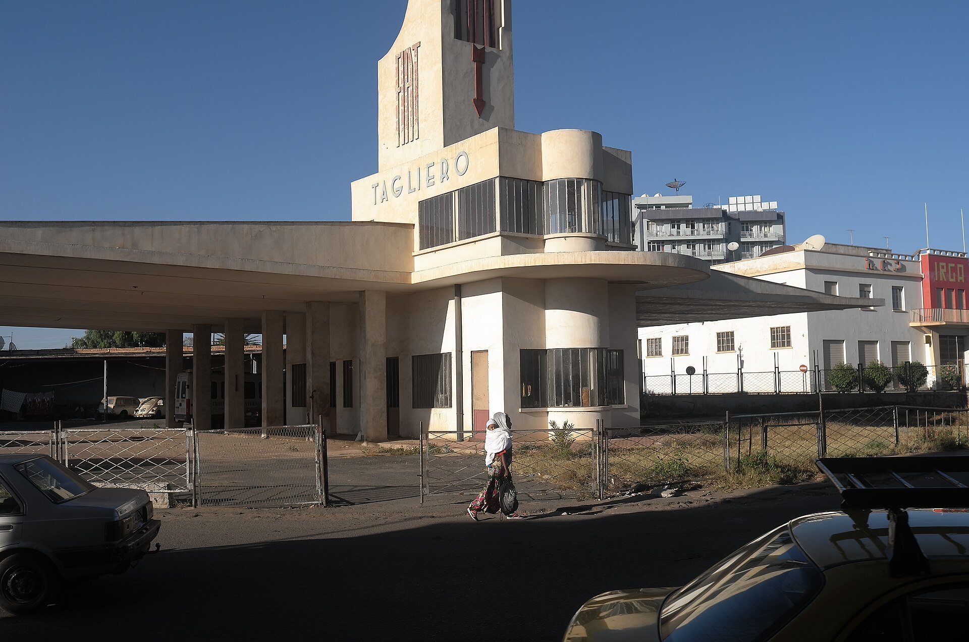 A white modernist petrol station in Asmara with a tall tower and a wide, flat canopy, a pedestrian passing in the street in front.