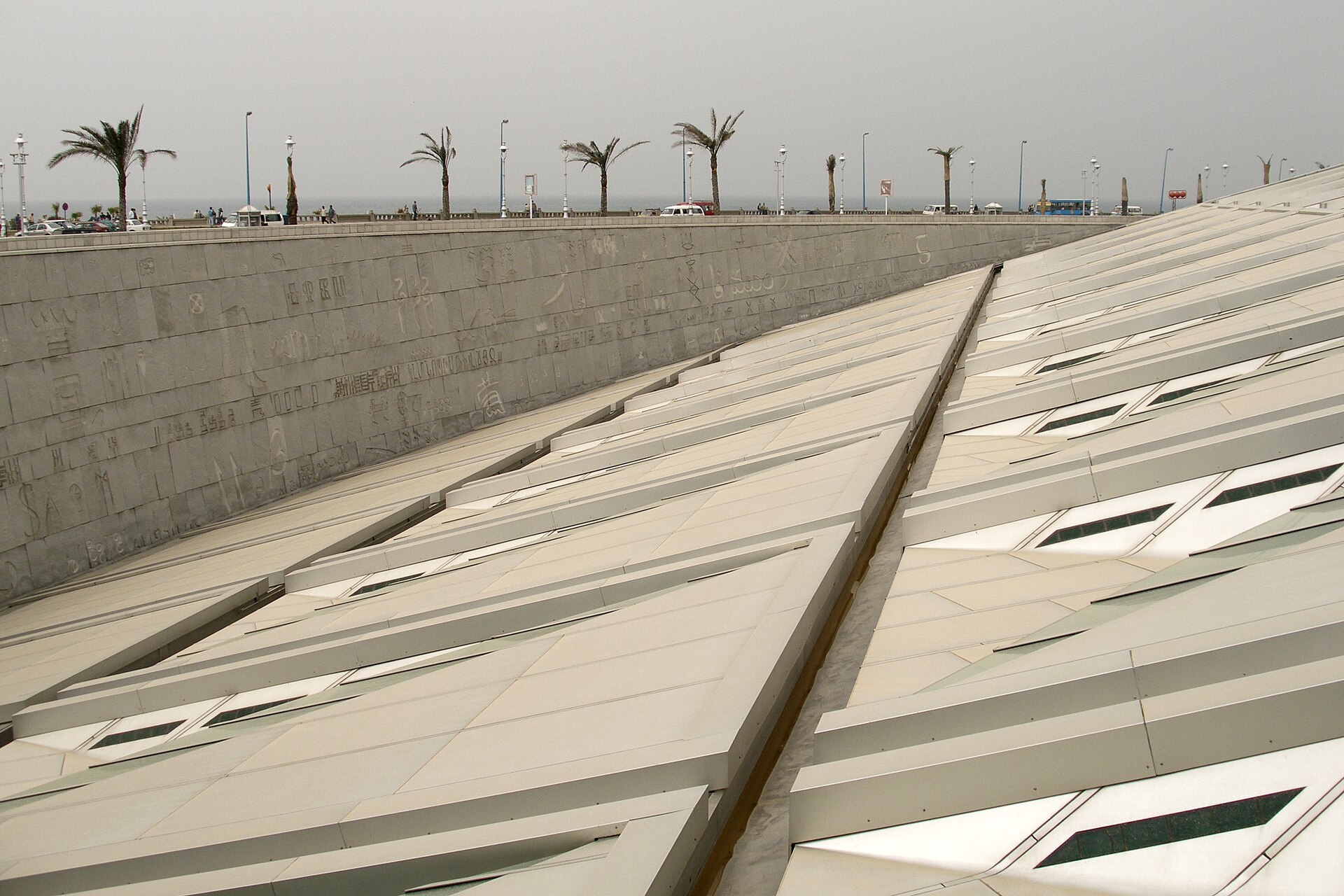 The tilted roof of the Bibliotheca Alexandrina with its skylights, beside the granite wall carved with signs, and palm trees along the sea front.