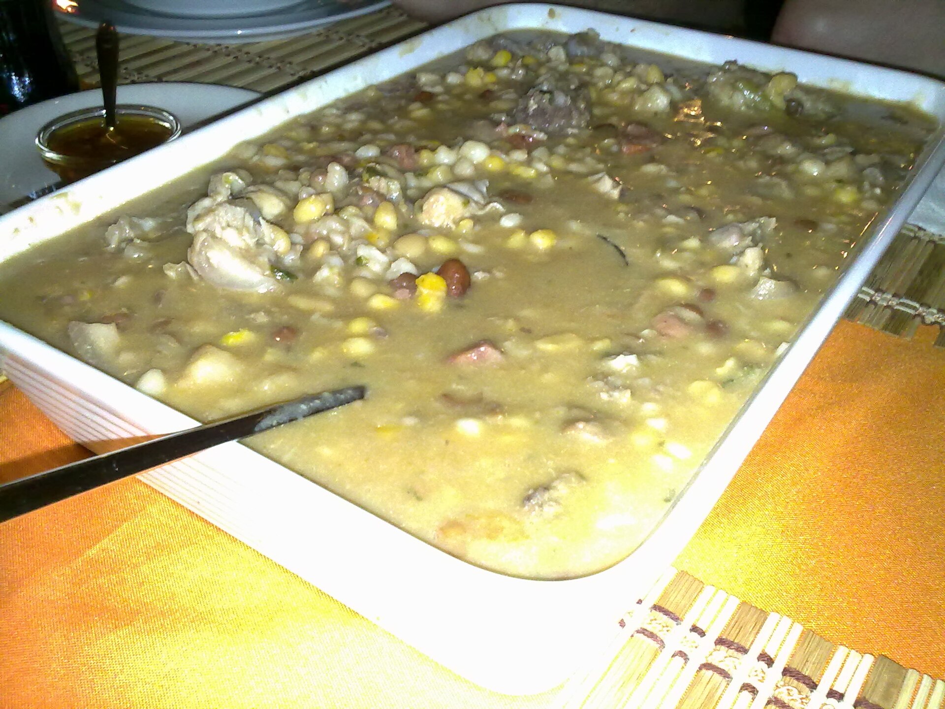 A large white dish of cachupa, a pale stew with corn kernels, beans and pieces of meat, with a ladle resting in it.