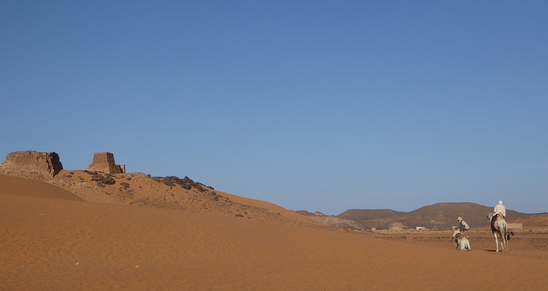 Small ruined pyramids on a ridge of orange desert sand, with two people and camels in the foreground.