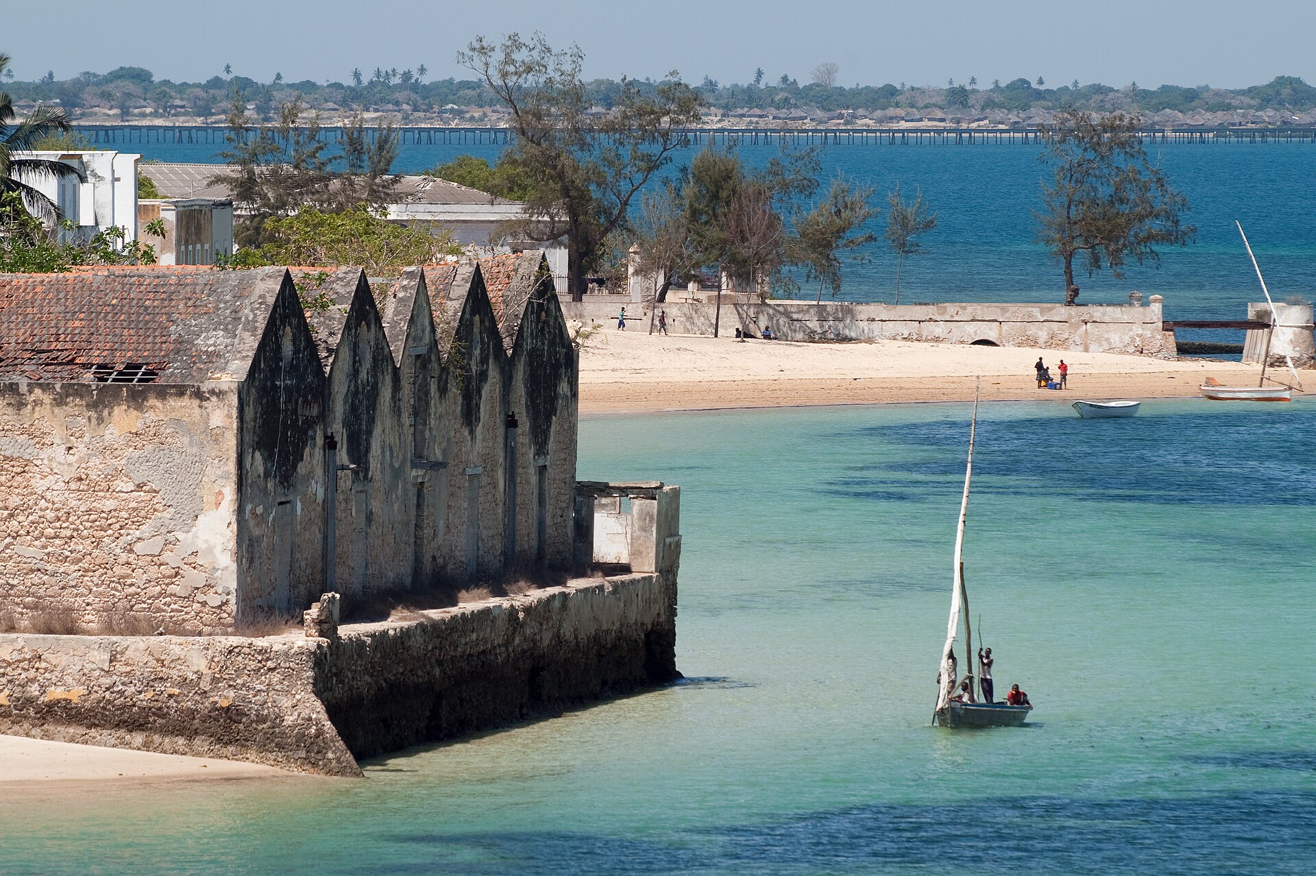 A small stone building with pointed gables on the shore, and a white sailing boat in turquoise water.