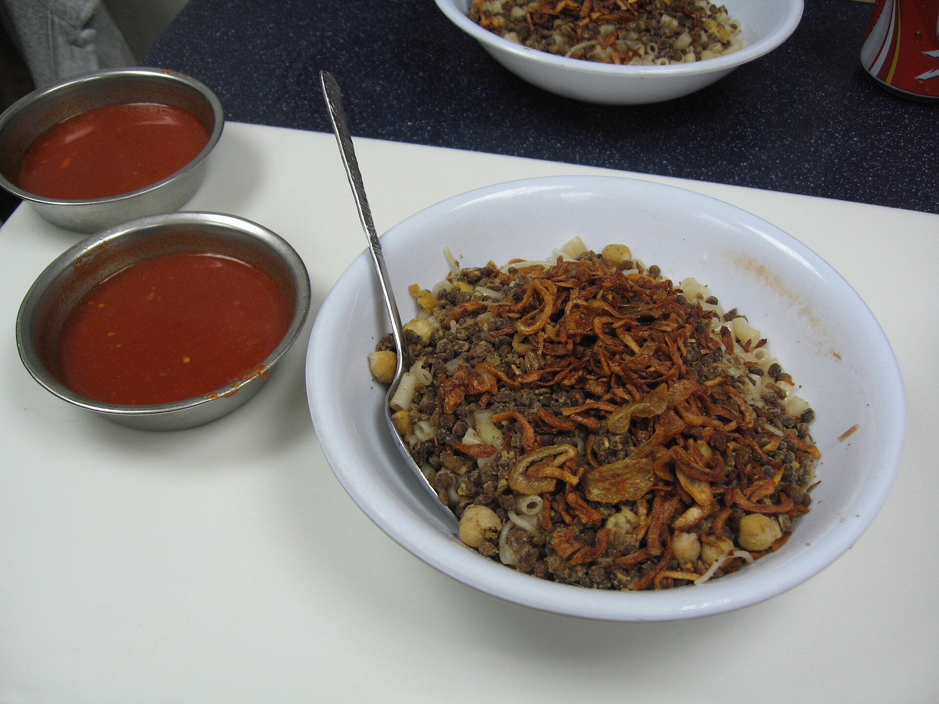 A white bowl of koshari piled with fried onions and chickpeas, with a spoon in it and two metal bowls of red sauce beside it.