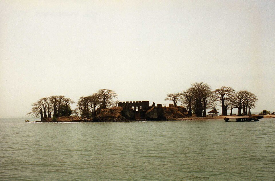 A low island in a wide, hazy river, with the ruins of a fort among bare baobab trees and a wooden jetty on the right.