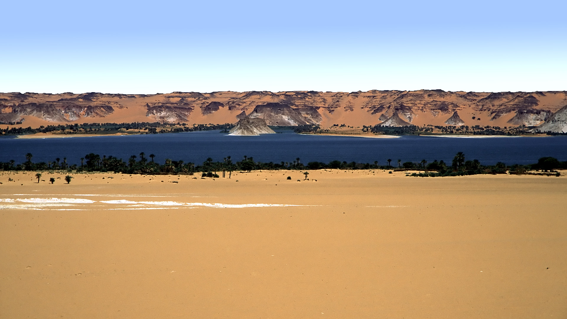 A still lake in the desert, with a strip of yellow sand in front and rocky cliffs behind.