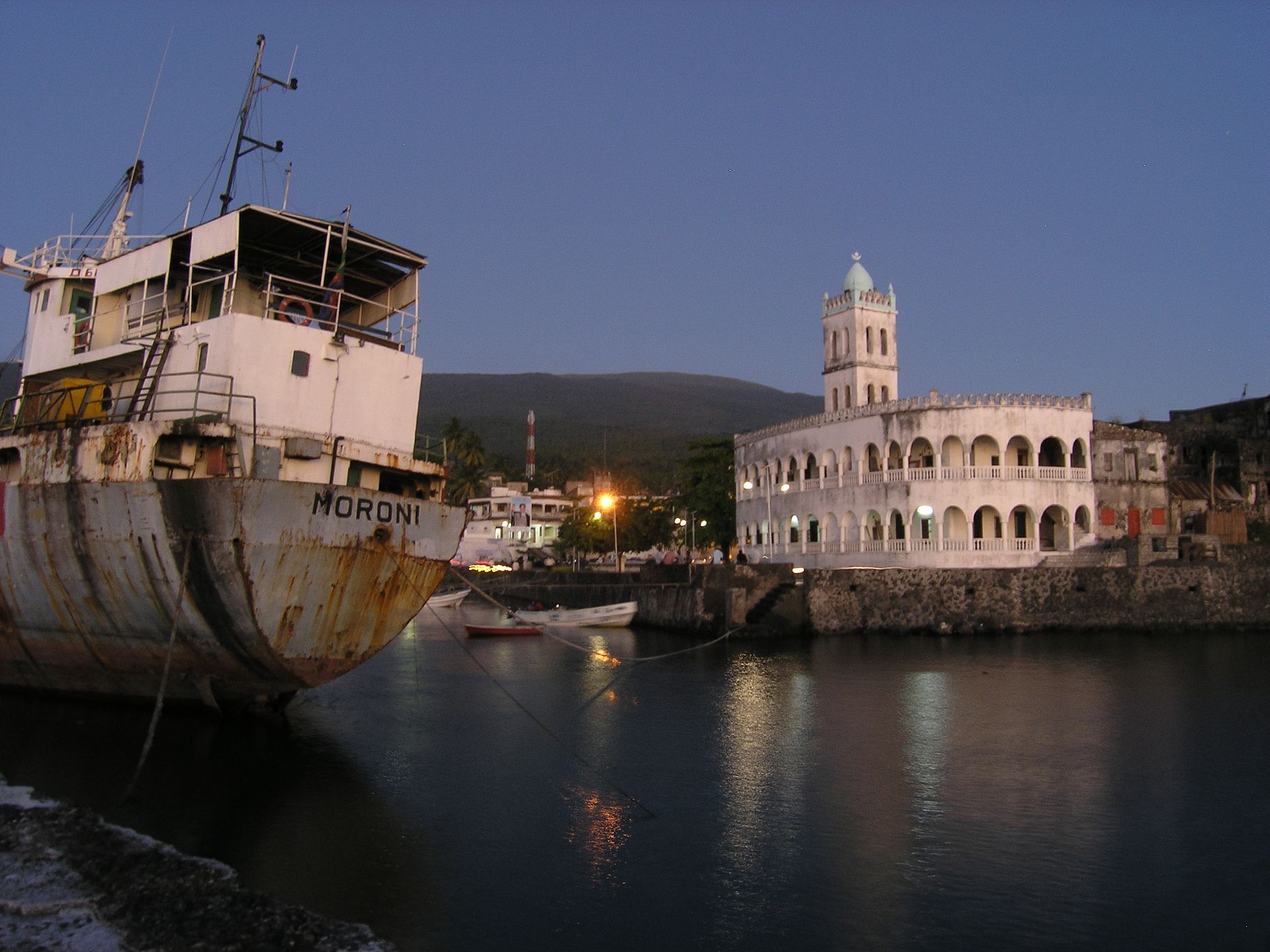 A white arcaded mosque with a small tower beside the harbour at dusk, a rusting ship moored on the left.
