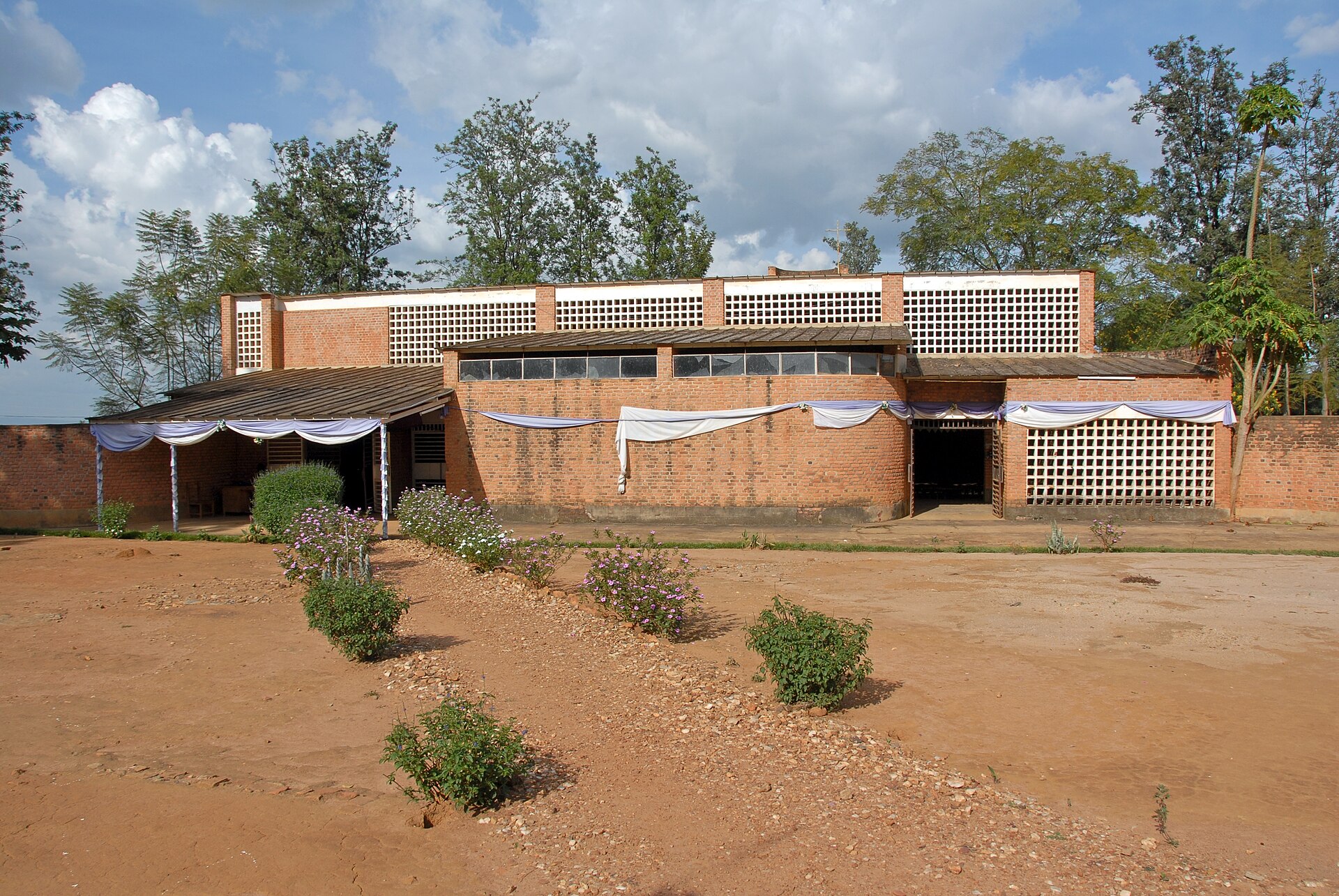 A low brick building with a long, flat roofline and ribbed walls, set back across a red-earth yard with young shrubs.