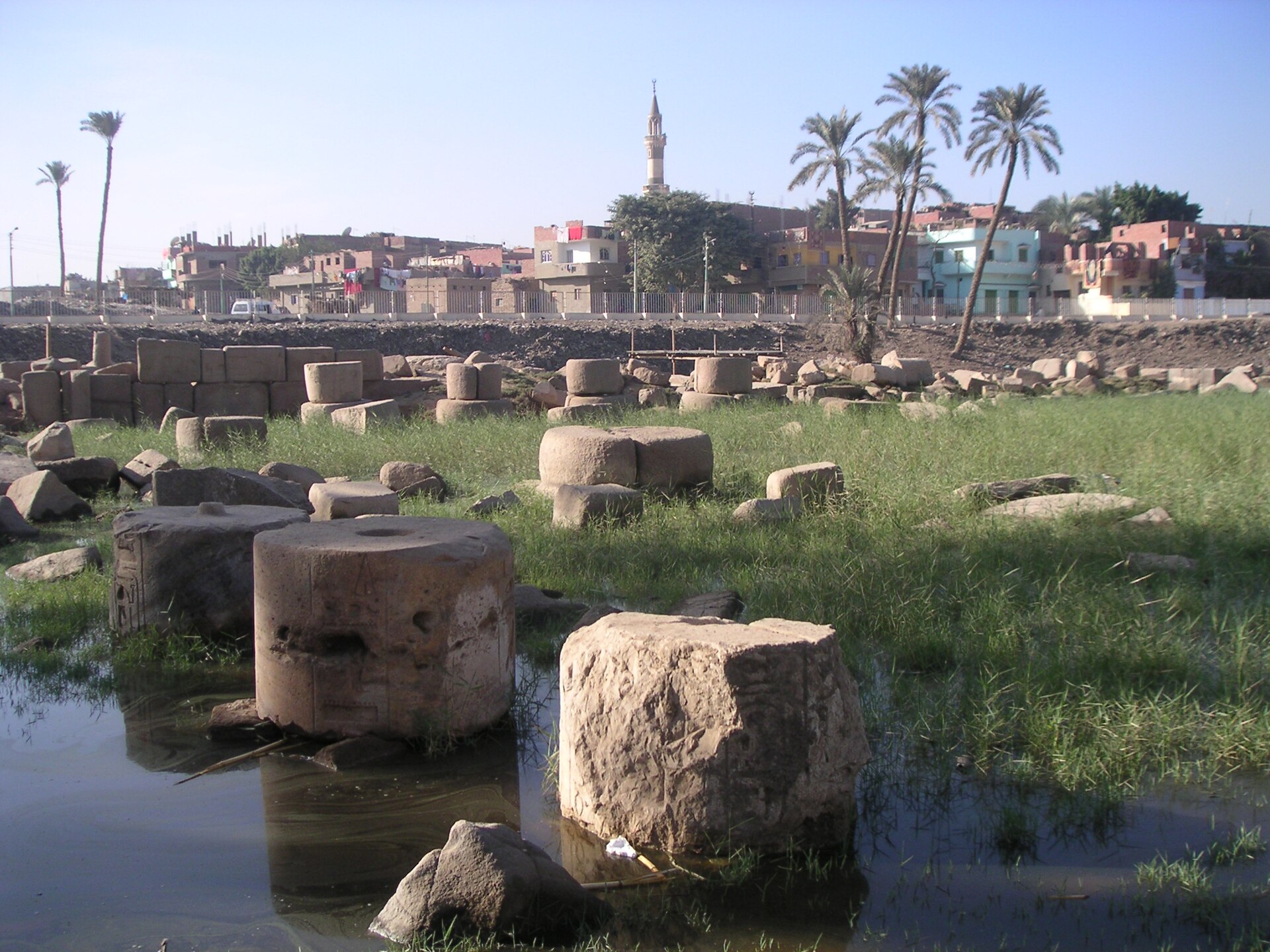Fallen stone column drums in grass and shallow water, with houses in the distance.
