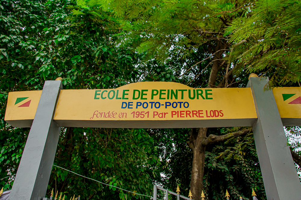 A yellow sign over a gate under trees, reading Ecole de peinture de Poto-Poto, founded in 1951 by Pierre Lods.