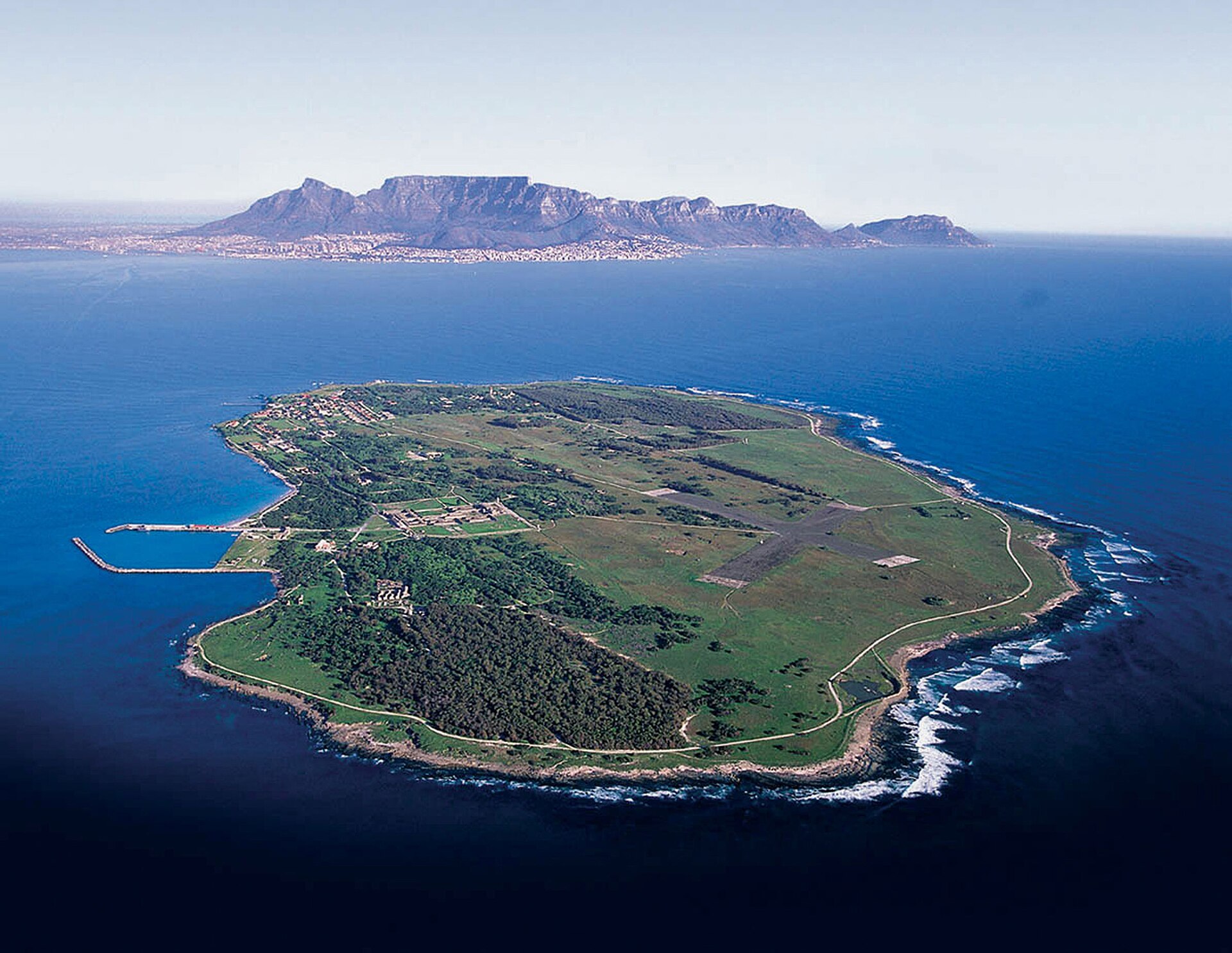 An aerial view of Robben Island, flat and green with a small harbour, in blue sea with Table Mountain on the horizon.