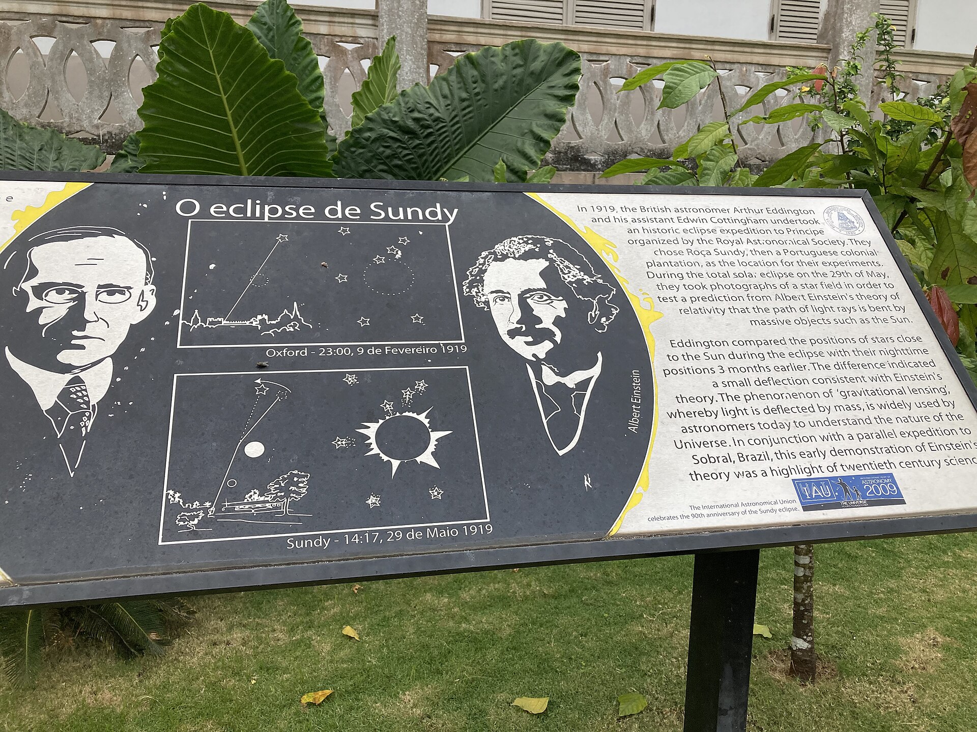 An information panel at Roça Sundy with portraits of Eddington and Einstein and diagrams of the 1919 eclipse photographs, standing on grass.