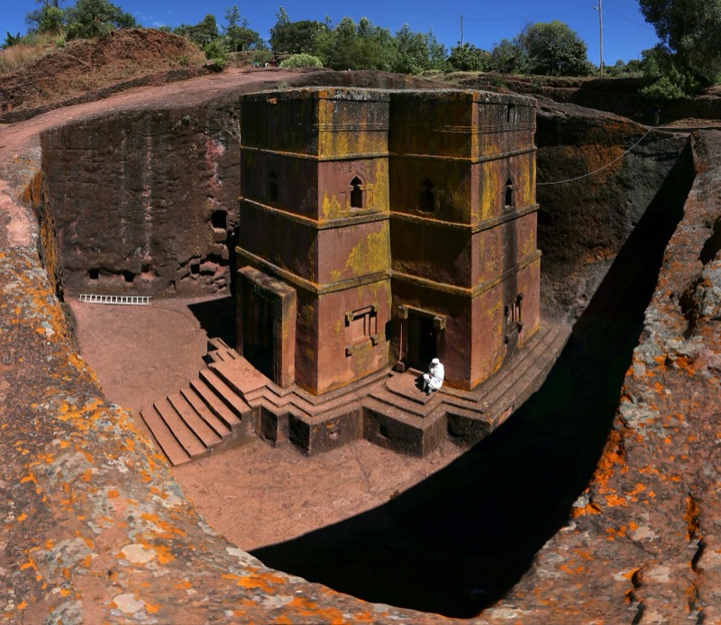 A rock-hewn church: a block of reddish stone standing in a pit, with steps leading down to its door.