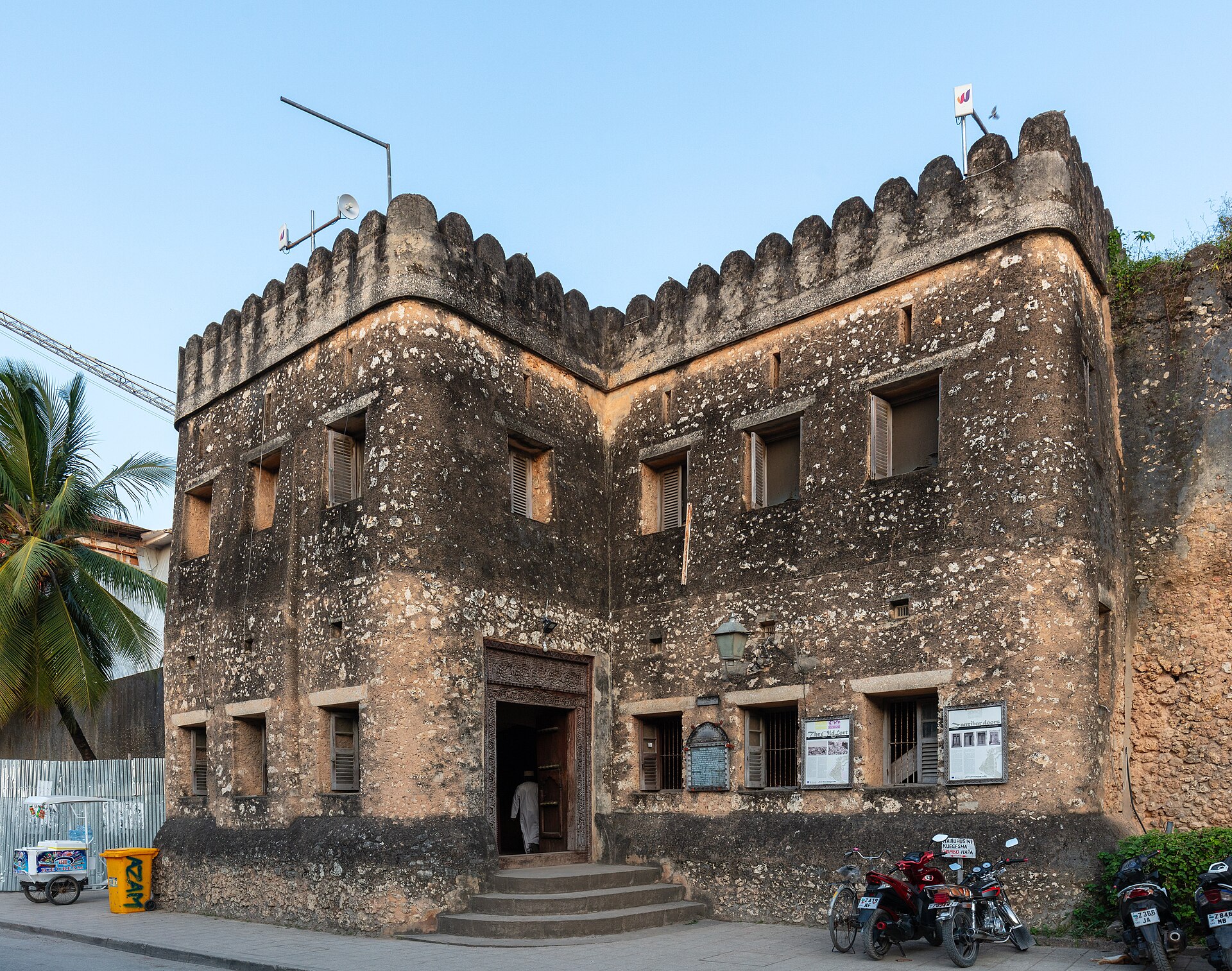 A weathered stone fort with crenellated towers and a carved doorway, palm trees and parked motorbikes in front.