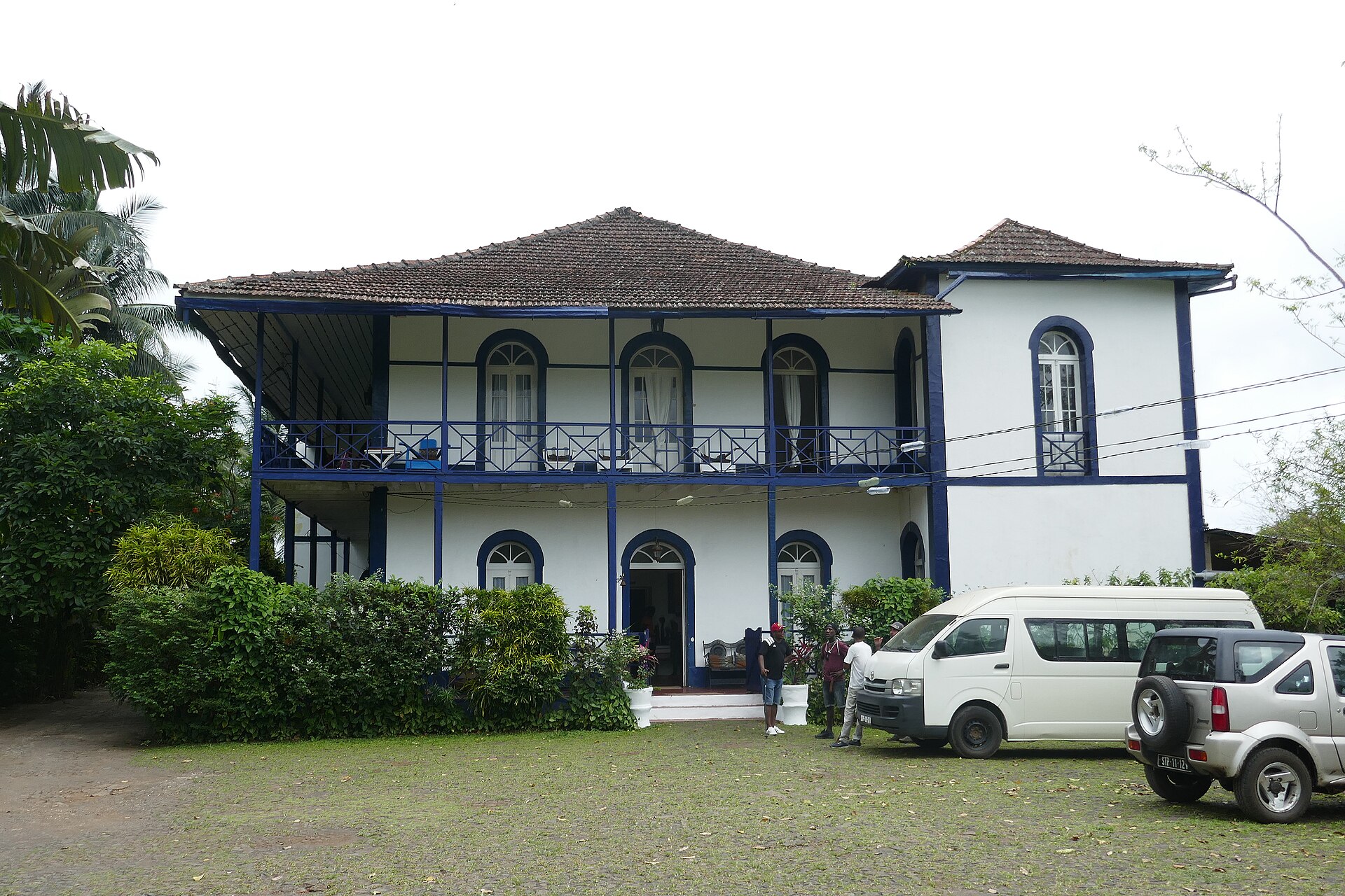 A two-storey white colonial house with a blue-trimmed veranda and arched windows, vehicles parked on the lawn.