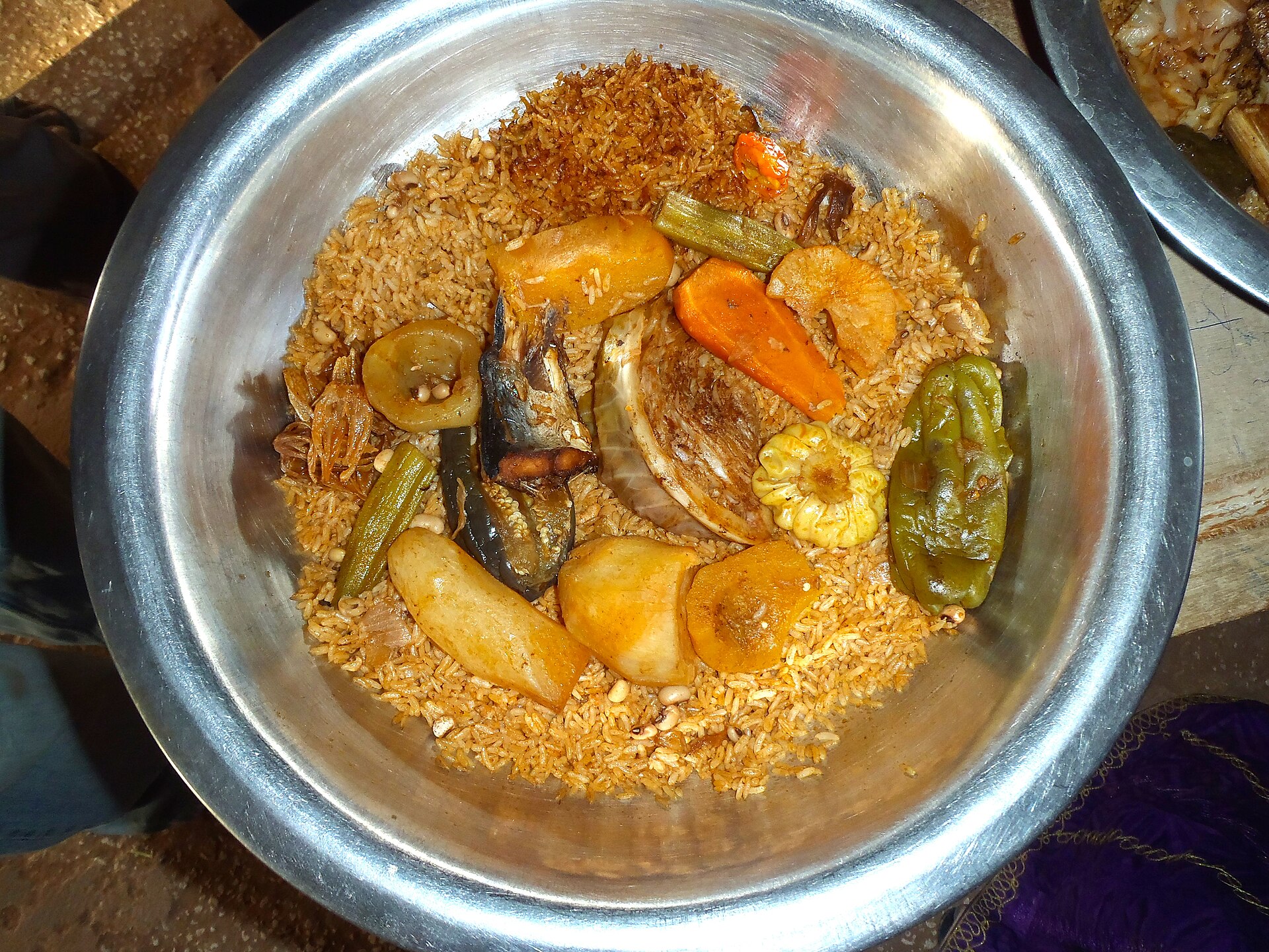 A large steel bowl of orange-brown rice with whole pieces of fish, carrot, cassava, aubergine and chilli laid on top.