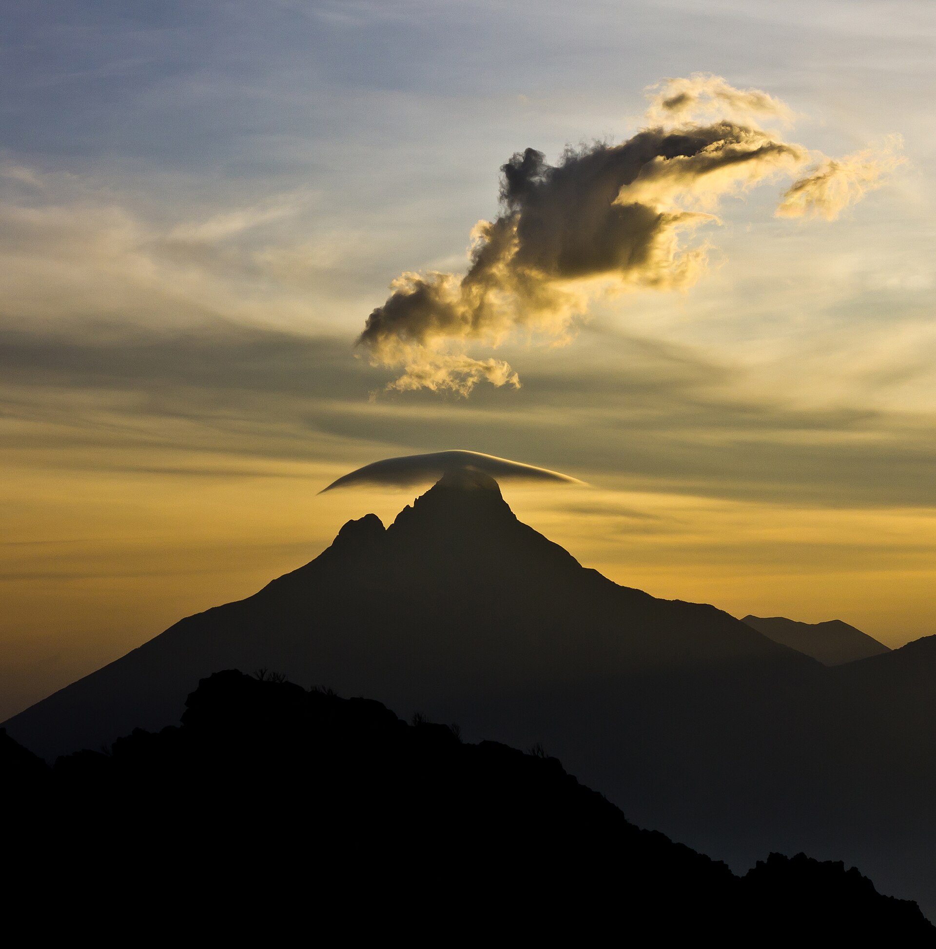 The dark silhouette of a volcanic peak at dusk, a small cloud above its summit lit gold by the setting sun.